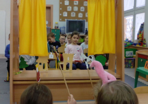Dzieci siedzące na krzesłach obserwują twórcze przedstawienie lalkowe dwóch koleżanek.